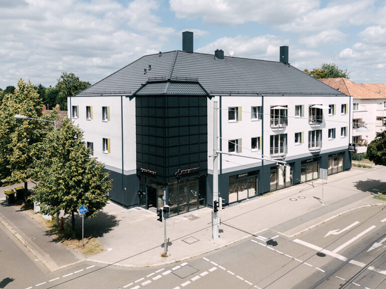 Außenaufnahme des CUBE Store Braunschweig. Modernes, dreistöckiges Gebäude an einer Straßenecke mit weiß-grauer Fassade, Geschäften im Erdgeschoss und Wohnungen mit Balkonen darüber. Im Vordergrund eine Kreuzung mit Ampeln und Zebrastreifen.